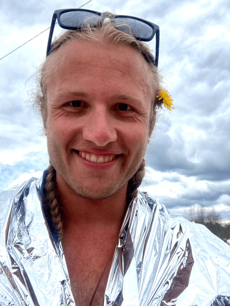 Mac, smiling, wearing a space blanket and with a dandelion behind his left ear.